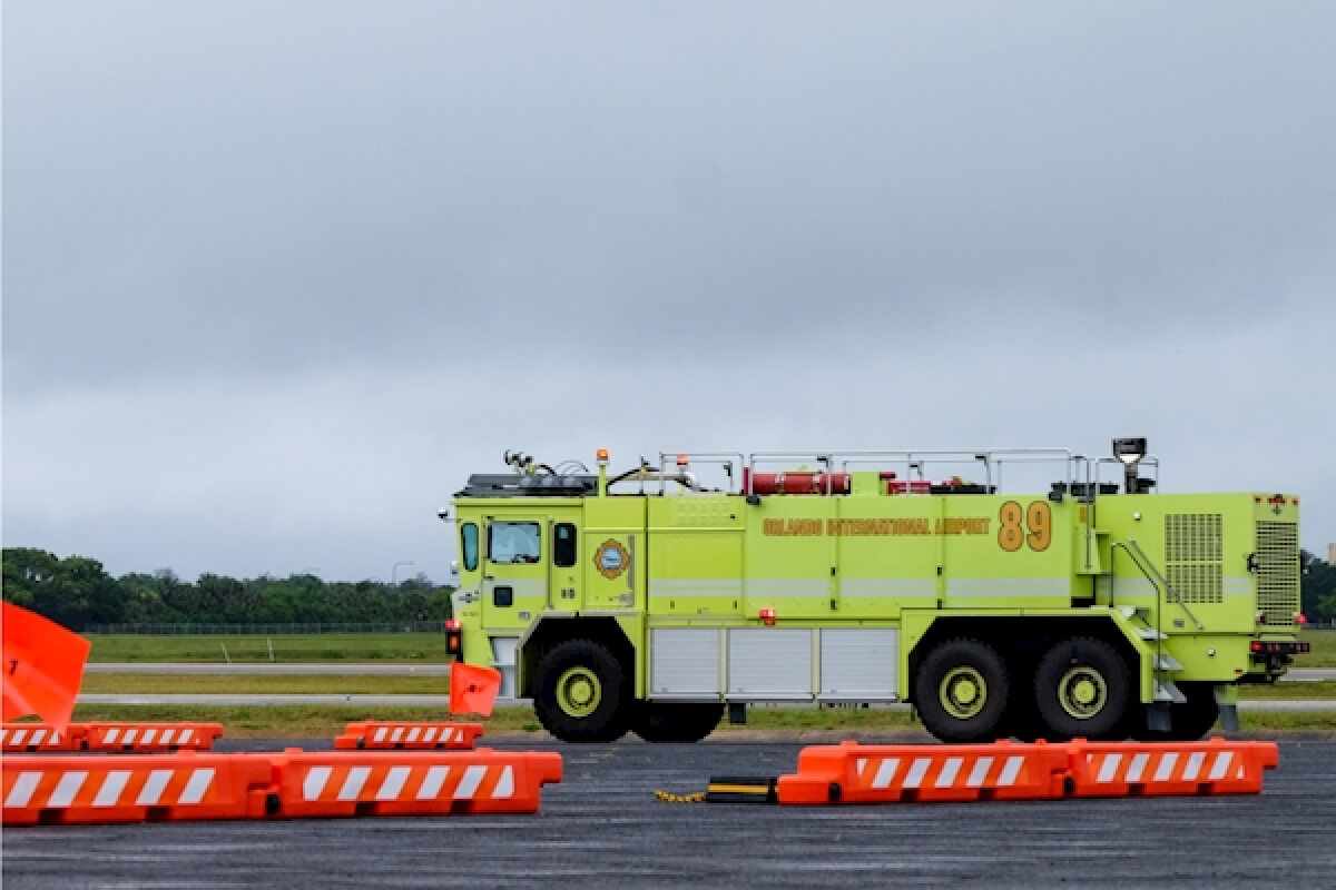 Airport fire truck parked on a wet runway near bright orange barricades under a cloudy sky.